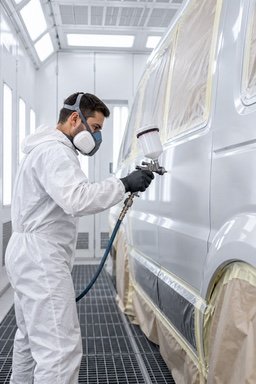 A painter in a protective suit and respirator spraying a masked van in a paint booth.
