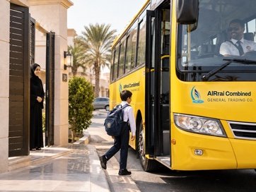 A boy with a backpack walks from his home gate to a waiting yellow AlRrai Combined school bus while his mother watches from the door.